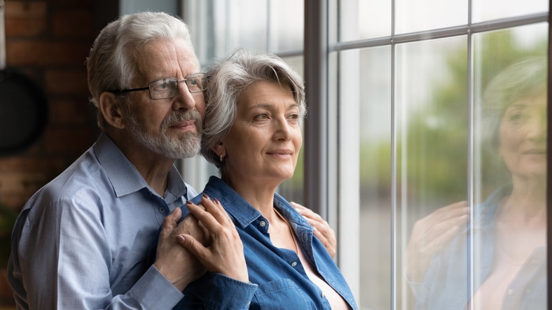 An older woman who is one of many experiencing anxiety in seniors is comforted by her husband as they gaze out the window together.