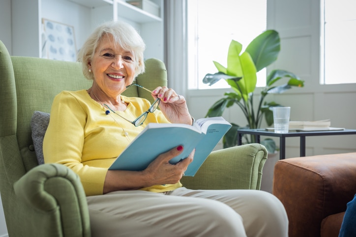 An older woman who is aging in place at home relaxes in her favorite chair with a book.