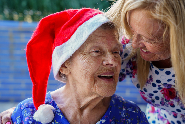 An older woman wears a Santa hat and laughs as her daughter shares holiday activities for older adults with her.